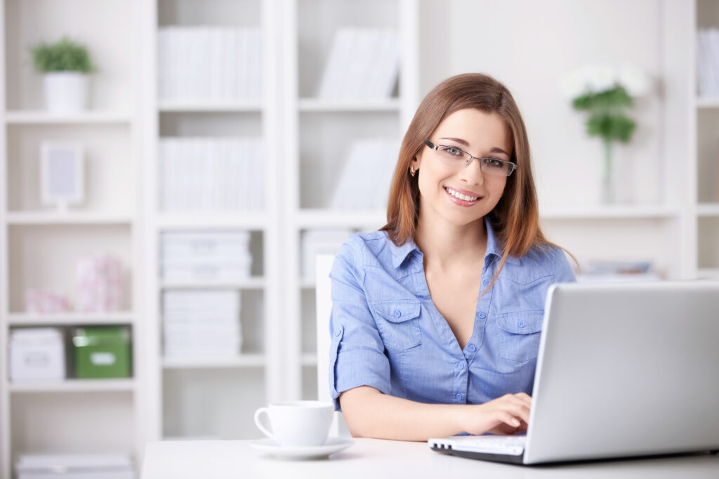Smiling young woman in a blue shirt works on a laptop at a modern desk, with a coffee cup and bookshelves in the background, suggesting a productive workspace.