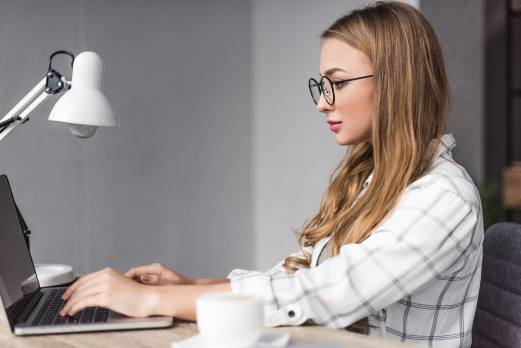 Woman with long hair and glasses types on a laptop at a wooden desk, with a desk lamp and a cup of coffee nearby, in a modern workspace.