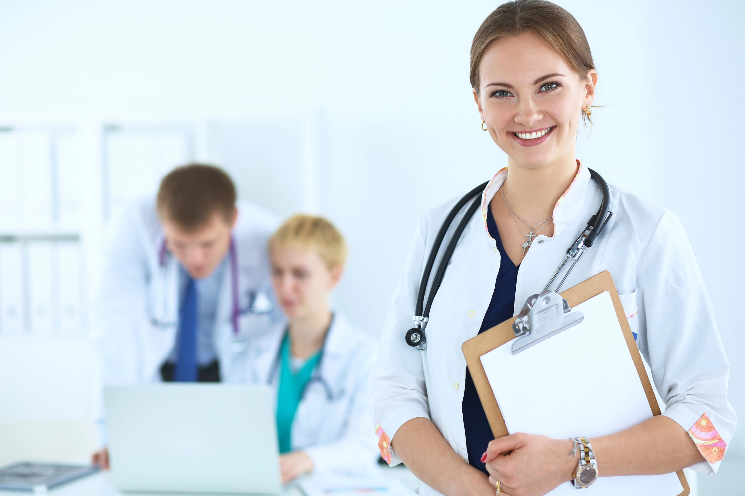 A confident female doctor smiles while holding a clipboard, with two colleagues in the background discussing medical information.