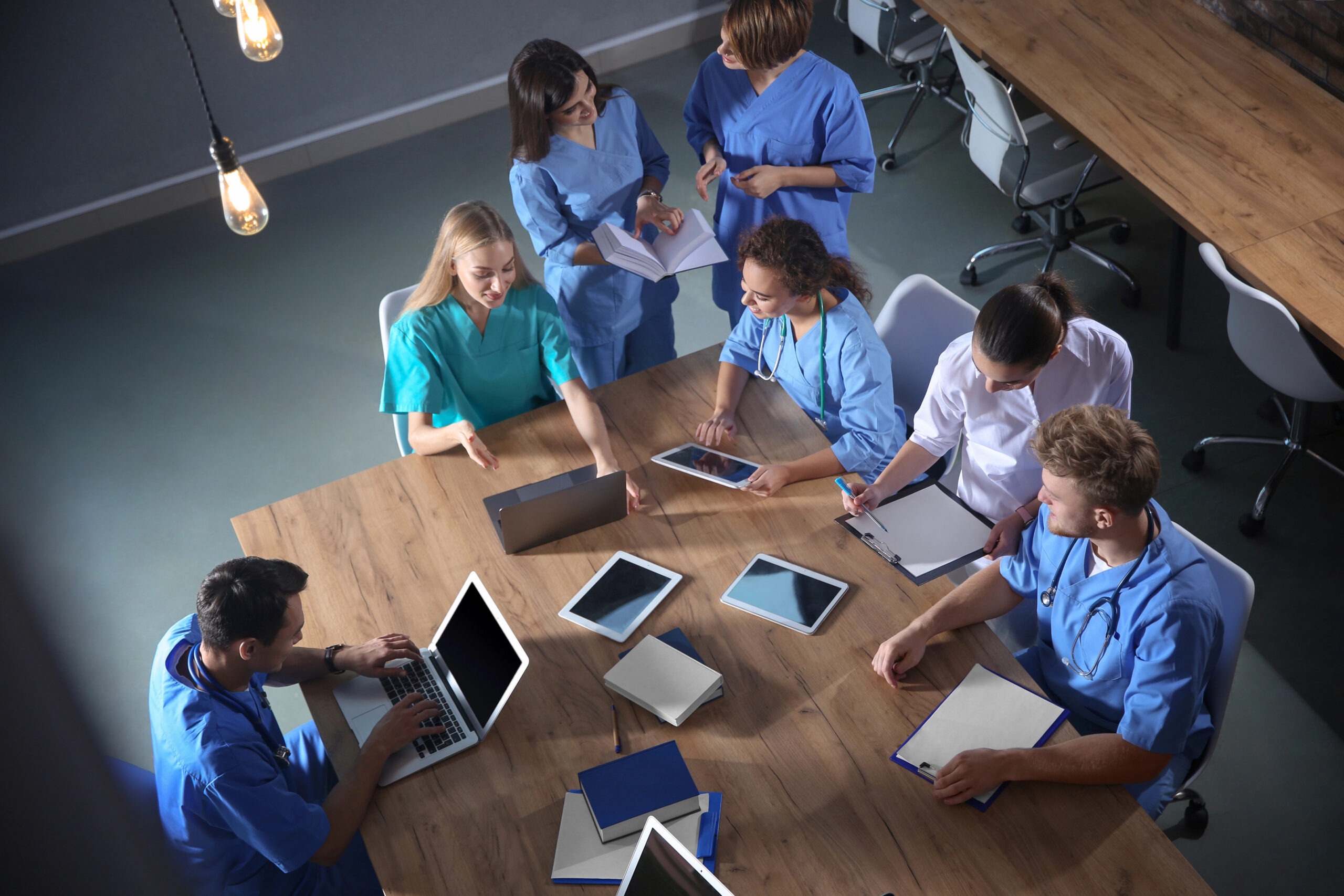 Healthcare professionals in scrubs collaborate around a conference table, engaging with laptops and tablets during a team meeting.