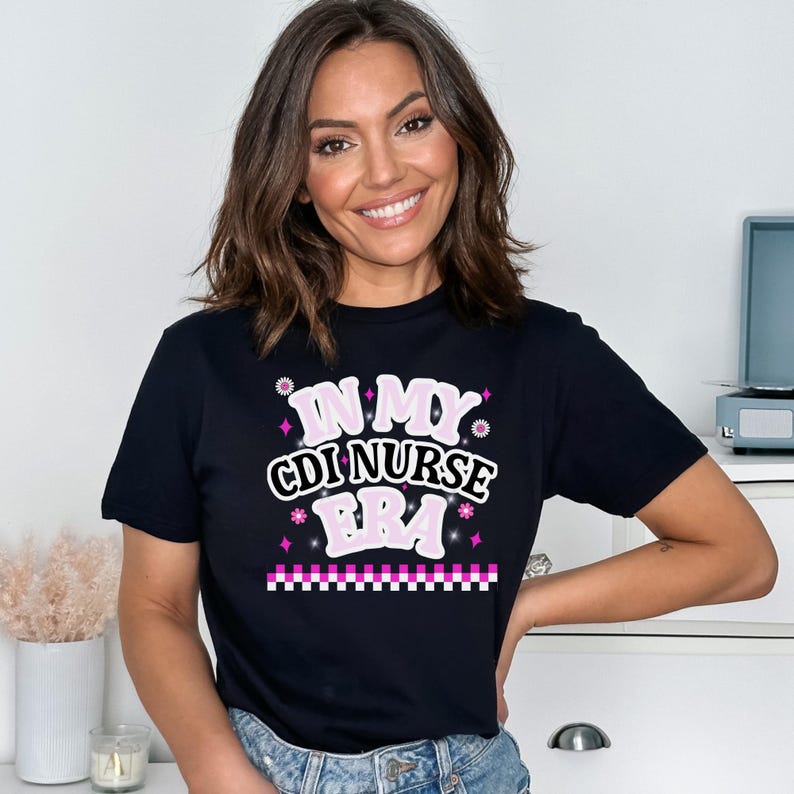 Woman wearing a black t-shirt with "IN MY CDI NURSE ERA" graphic, smiling against a neutral background with decor items. Celebrates nursing identity.