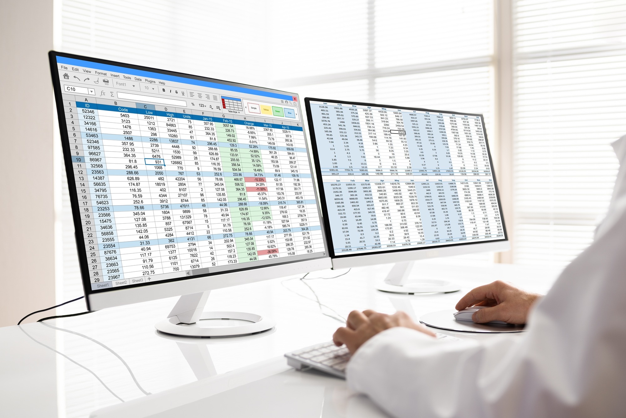 A person analyzes data on dual monitors displaying spreadsheets with various financial metrics and calculations. The setting suggests a professional workspace.