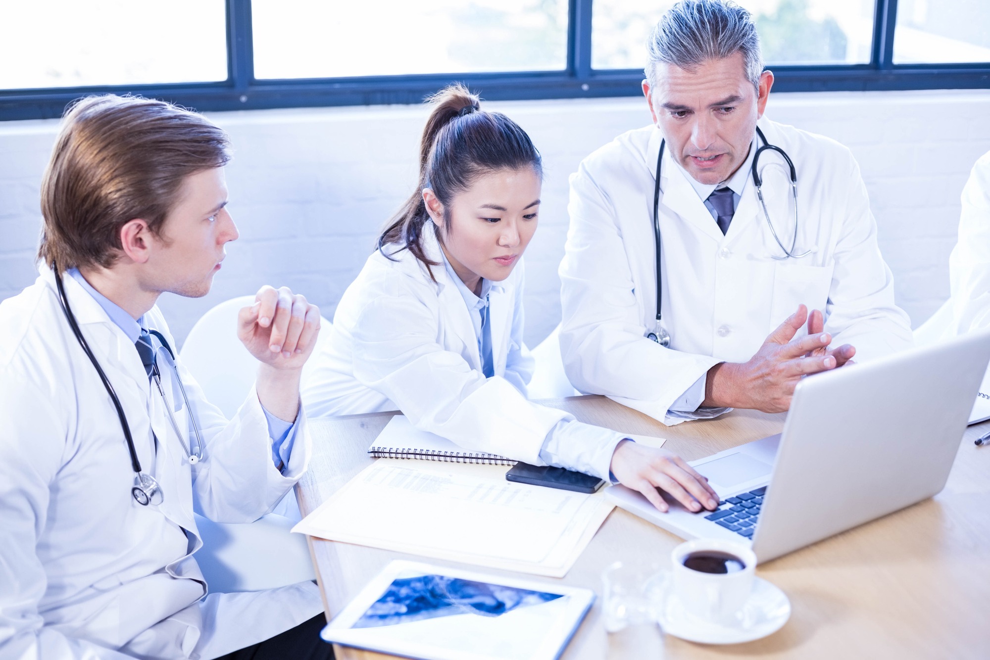Three doctors in white lab coats collaborate at a table, discussing a laptop and reviewing documents while engaged in conversation.